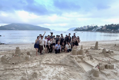 A large group of people posing together on a sandy beach behind several detailed sandcastles they built, with the sea and mountains visible in the background under an overcast sky.