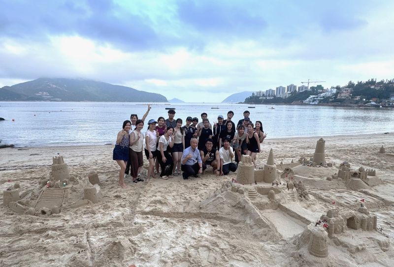  A large group of people posing together on a sandy beach behind several detailed sandcastles they built, with the sea and mountains visible in the background under an overcast sky. 