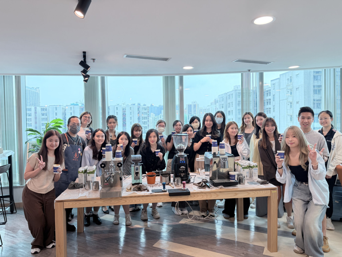 A large group of people posing together around a wooden coffee bar setup indoors, with large windows overlooking city buildings in the background.
